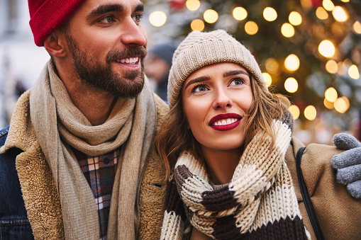 Smiling man is hugging beloved girlfriend while they are walking in city centre during winter holidays