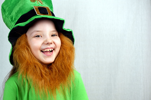 Portrait of adorable laughting seven years old girl with decorative red beard in green clothes and leprechaun's hat. Saint Patricks Day celebration