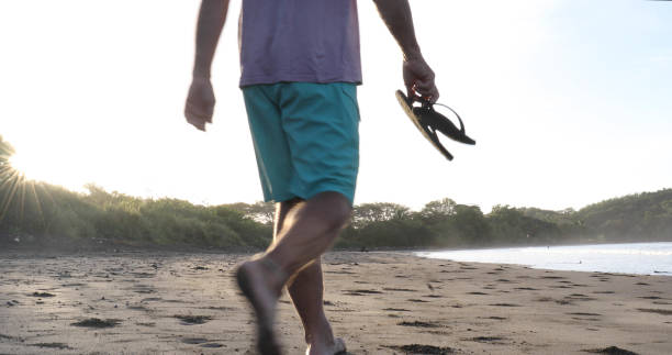 istock Man holding sandals walks down sandy beach at sunrise 1299814887