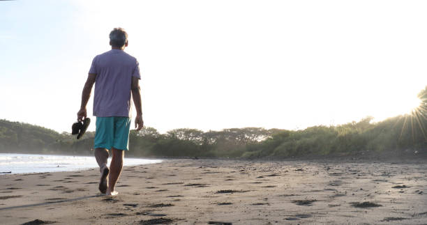 istock Man holding sandals walks down sandy beach at sunrise 1299815428