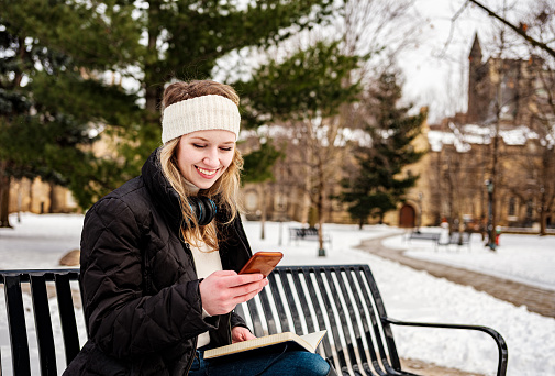 Female university student reading her notes while sitting on the park bench. Exterior of city public park in winter  with historic buildings in the background.