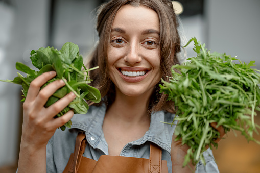 Portrait of cheerful woman in apron with fresh spinach and arugula on the kitchen. Healthy cooking concept. Close up. High quality photo