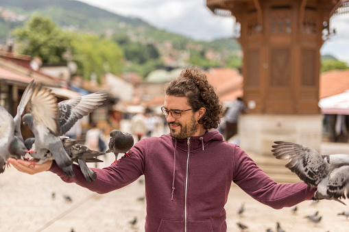 Handsome young man having fun feeding flock of pigeons with seeds on city square while on vacation, holding arms spread with numerous pigeons eating from his hands