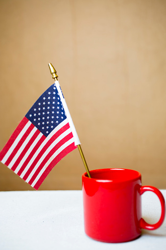 Small American flag sits in a red ceramic mug on a white top table.  Solid wall in background for copy space.