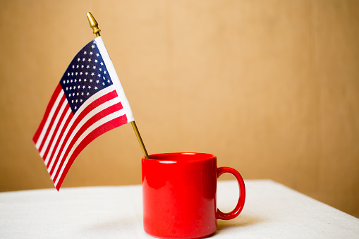 Small American flag sits in a red ceramic mug on a white top table.  Solid wall in background for copy space.