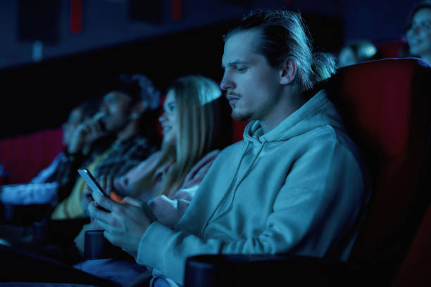 istock Focused young guy using his smartphone while missing boring movie at the cinema 1299842009