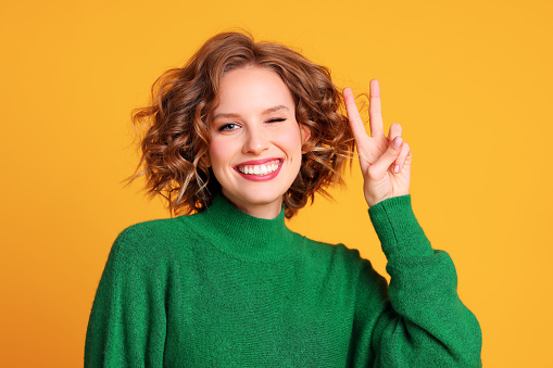 Delighted young woman in stylish green sweater winking at camera and showing V sign against yellow background