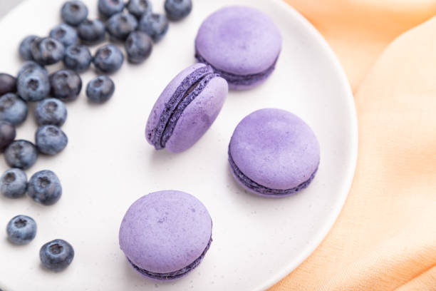 istock Purple macarons or macaroons cakes with blueberries on white ceramic plate on a gray concrete background and orange textile. Top view, close up, selective focus. 1299847269