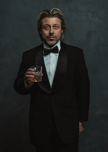 Satisfied blonde man with stubble beard in tuxedo holding glass of whisky.