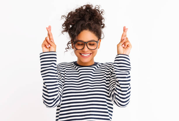 Happy young ethnic female making wish Cheerful young ethnic woman in glasses closing eyes and crossing fingers while making wish against grey background good luck stock pictures, royalty-free photos & images