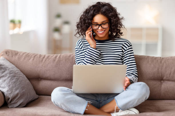 mulher étnica alegre com laptop falando no smartphone - 15821 - fotografias e filmes do acervo