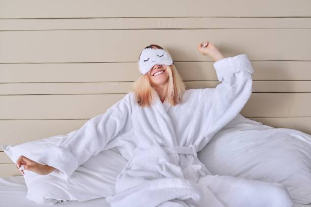 młoda kobieta w białym łóżku w szlafroku, masce na oczy, żeńskich ziewa - pillow wake up yawning sleeping zdjęcia i obrazy z banku zdjęć