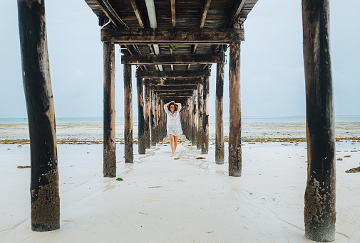 Portrait of a happy cheerful smiling woman dressed in light summer clothes walking barefoot under the sandy beach wooden pier. Careless vacation in the tropical countries concept image