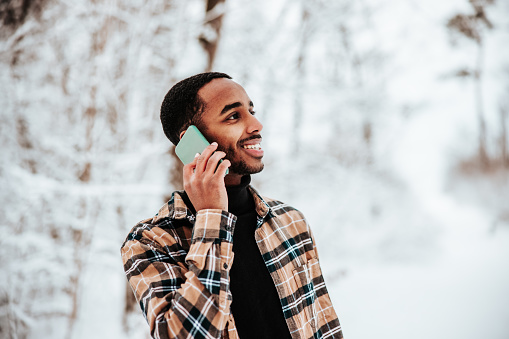 Happy young fashionable black man using his mobile phone for a telephone call while standing outdoors in winter with a bright happy smile.Talking, laughing and having a good chat. Millennial Generation Real People Outdoor Lifestyle Portrait.