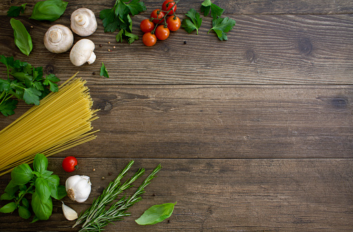 A selection of Italian ingredients and herbs on a rustic wooden  background. \nGarlic, basil, mushrooms, spaghetti, tomatoes, rosemary and dried peppercorns. \nShot from above with copy space on the right of the image. Italian food/ cooking flat lay.