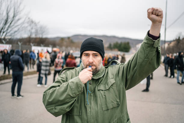 istock Young man with raised fist protesting on a street blowing a whistle 1299865485