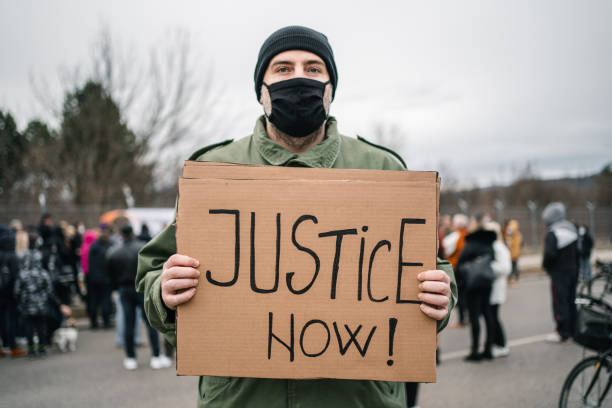 istock Young man with protective mask holding a banner with text Justice now 1299865507