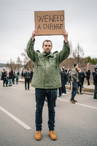 Young angry man protesting on a street holding a placard with text We need a change with large crowd of people