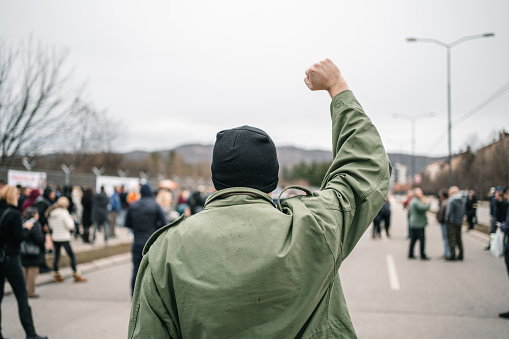 Young angry man with raised fist protesting on a street blowing a whistle with large crowd of people