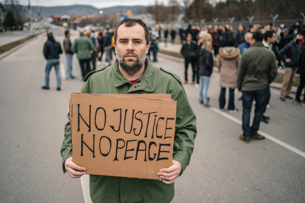 istock Angry man protesting on a street holding a sign with text No justice no peace 1299865679