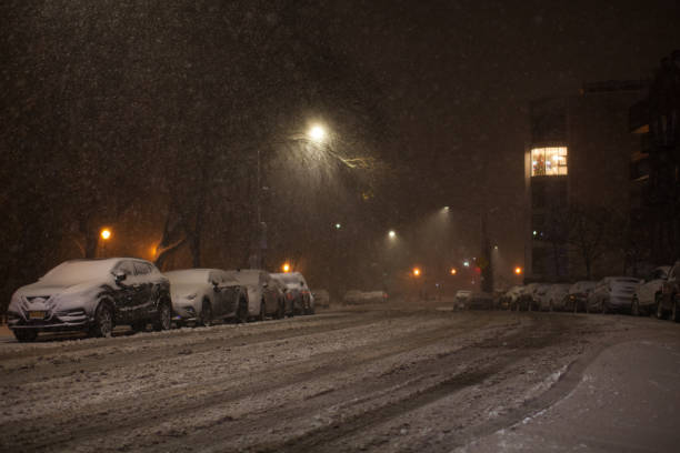 The photo shows a snow-covered road in a residential area of Brooklyn. There are traces of cars are visible on the snow. The sidewalk is not cleared of snow at all. It's very hard to walk. Winter storm in progress and snow gathers in drifts. Along the edges of the photo are two-story brick houses. The branches of the trees growing along the edges of the road. Trees and branches completely covered with snow. Cars parked at the sides of the street are also covered with snow. Snow continues to fall and snowflakes are visible flying in the air. Winter storm in progress and snow gathers in drifts. Night. December 16, 2020. NYC. Brooklyn. Bay Ridge. New York. USA