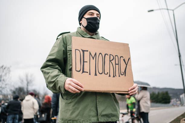 istock Protestor with protective mask holding a sign with text Democracy on a street 1299866392