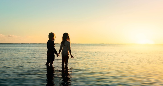 Two kids holding hands, standing in the ocean water watching the sunset. Future generation.