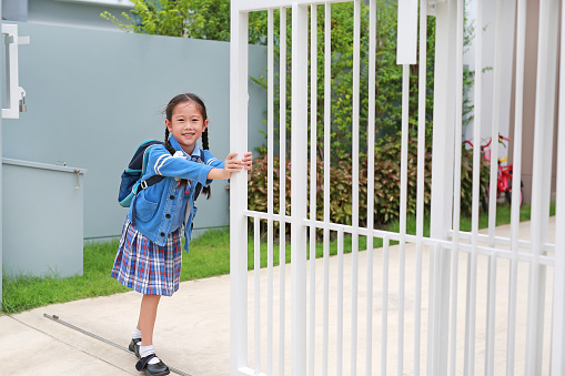 Portrait of happy asian little kid girl in school uniform trying push and pull door fence of house to close or open before leave home to go to school at morning.