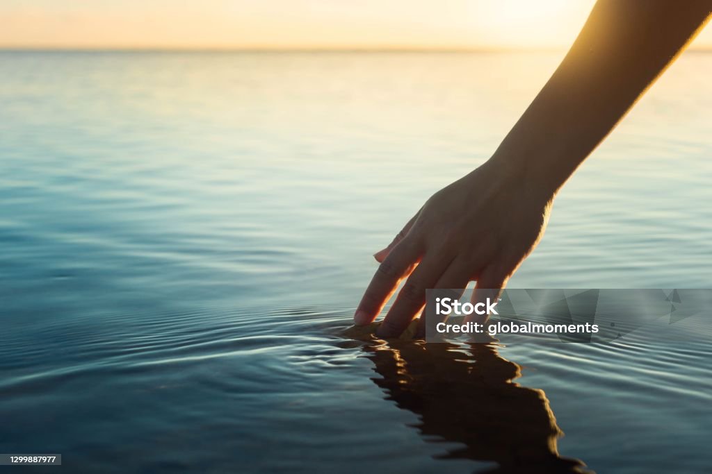 Happy people in nature. A woman feeling and touching the ocean water during sunset. A female hand touching the ocean water in front of a beautful sunset during summer time. Water Stock Photo
