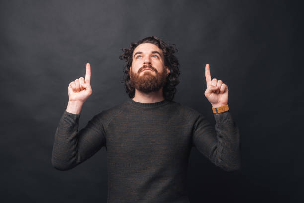 istock A young bearded man is looking up at a free space and at the same time is pointing with his fingers up at the same free space 1299895859