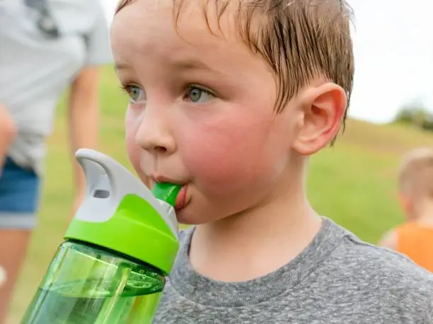 A young boy taking a break from vigorous exercise and playing outside, drinking water and sweating.