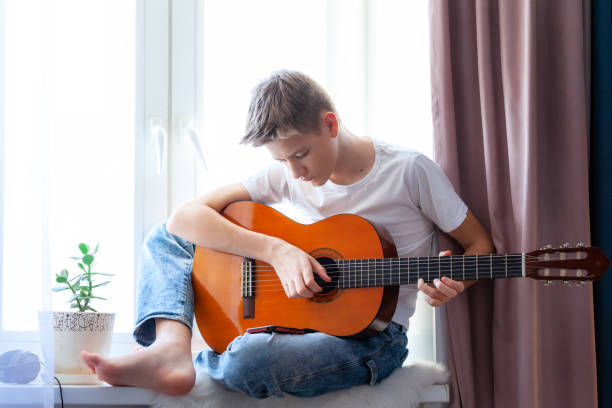 istock Handsome guy playing acoustic guitar while sitting on windowsill at home in his room 1299898756