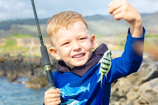 A young child holding a fish and a rode smiling and happy at the fish he caught near the ocean water.