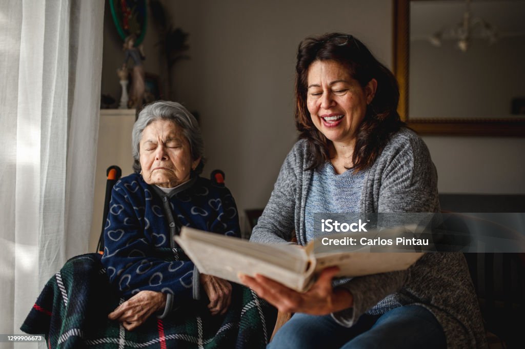 Old sick woman with memory loss sitting in wheelchair. Old sick woman with memory loss sitting in wheelchair. Smiling daughter holding a photo album trying to remember. Nursing Home Stock Photo