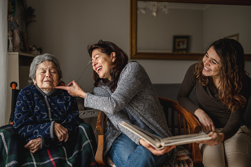 women generation with old sick grandmother sitting in wheelchair and smiling daughter and granddaughter looking a photo album.