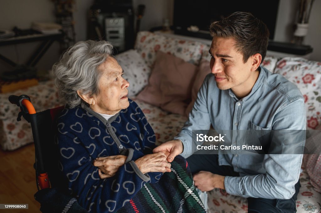 Young man sitting next to an old sick aged woman in wheelchair taking her hands while talking and smiling Young man sitting next to an old sick aged woman in wheelchair taking her hands while talking and smiling. Family, home care concept. Dementia Stock Photo