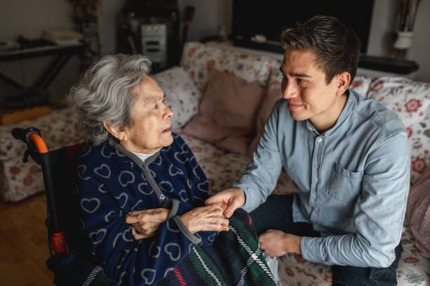 Young man sitting next to an old sick aged woman in wheelchair taking her hands while talking and smiling Young man sitting next to an old sick aged woman in wheelchair taking her hands while talking and smiling. Family, home care concept. dementia stock pictures, royalty-free photos & images