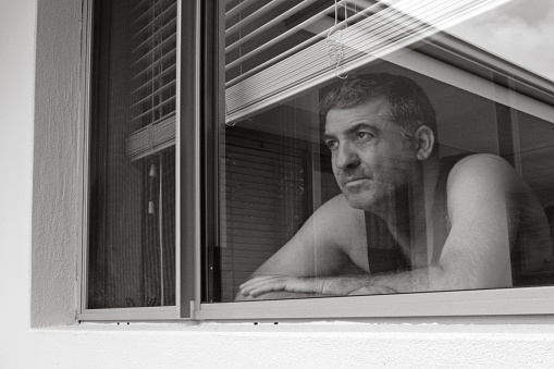 Sad adult man (age 40-50) looking out through home window.