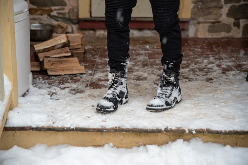 A young person in snow covered boots stands on snowy porch by a pile of freshly chopped firewood. Winter scene, yellow porch and stone house background.
