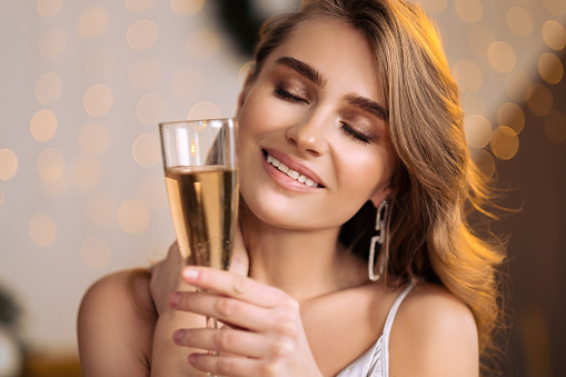 A young beautiful blonde woman in a silver cocktail dress holds a glass of champagne. Smiling and posing against the backdrop of a Christmas tree. Makes a wish and celebrates.