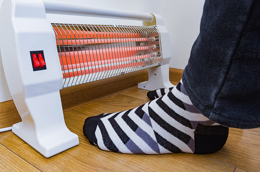 Person warming legs near an electric heater. Concept image of heating season at home