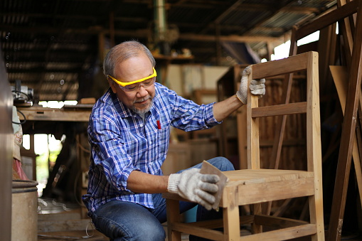 Senior Asian carpenter man is repairing wooden chair in his own garage style workshop at retirement age for hobby with copy space