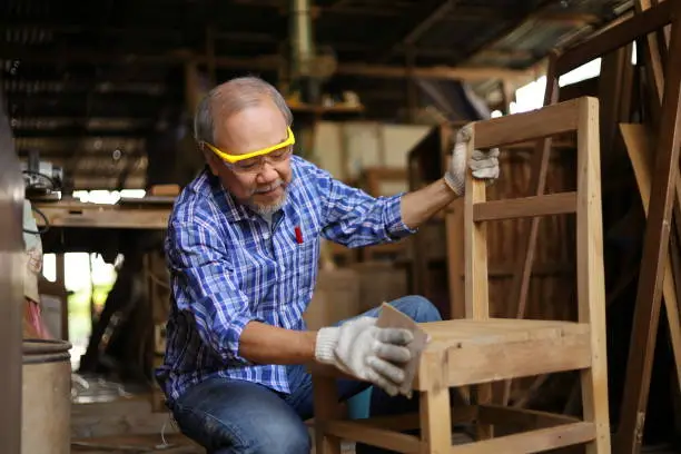 Photo of Senior Asian carpenter man is repairing wooden chair in his own garage style workshop at retirement age for hobby