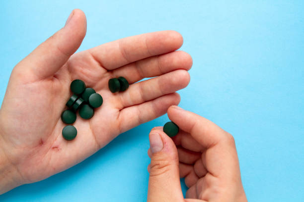 한 손에는 여러 개의 클로렐라 정제가 있습니다. 다른 손은 알 약 중 하나를 걸립니다. 파란색 배경입니다. 선택적 초점. - spirulina bacterium green vitamin pill medicine 뉴스 사진 이미지