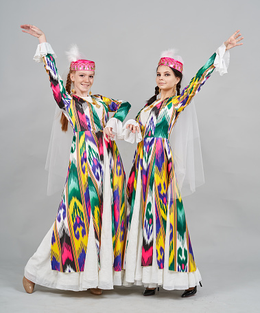 Two young caucasian woman are dressed in a Uzbek traditional clothing. The young females dancers are dancing national dance and showing traditional poses. They are smiling looking at the camera. Studio shooting on white background