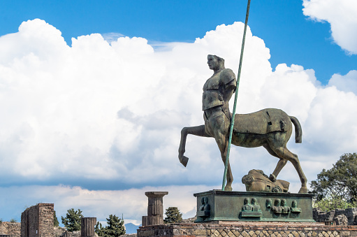 The bronze sculpture of Centaur (sculptor Igor Mitoraj) donated to Pompeii. Pompeii ruins in Italy, a background of collumns, trees and literally mind blowing clouds