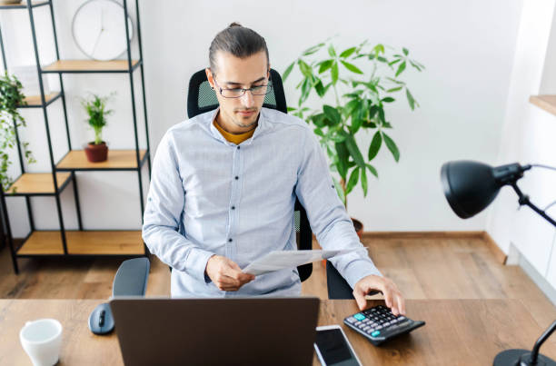 istock Man in office looking at a document and working on a laptop. / Remote working concept. 1299921032