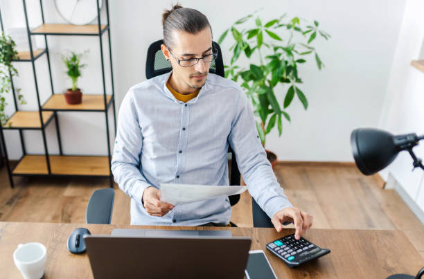 istock Man in office looking at a document and working on a laptop. / Remote working concept. 1299921033