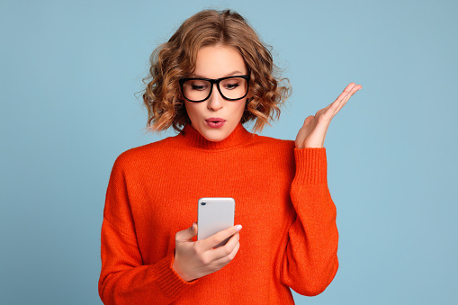 Amazed young female in orange sweater and trendy glasses reading unexpected message on smartphone   against blue background