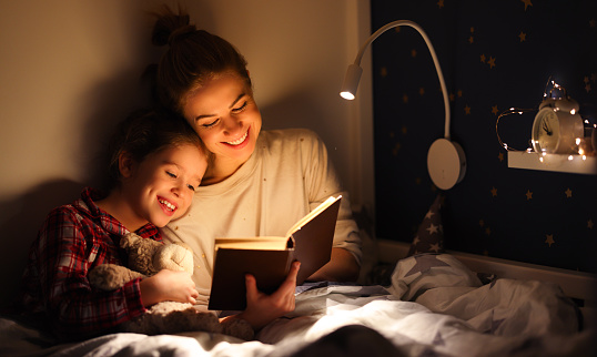 Cheerful family: woman and girl smiling and reading fairytale while relaxing on bed near lamp at night at home